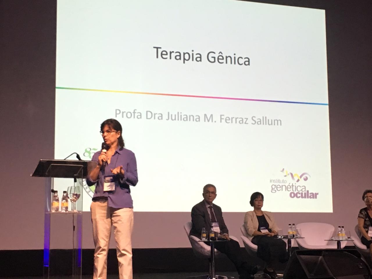 #PraCegoVer foto da Profa. Dra. Juliana Sallum palestrando no palco no evento. Ao fundo há uma projeção com o título "Terapia Gênica"