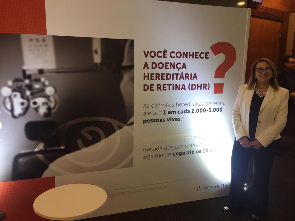 #PraCegoVer foto do evento. Na foto está a presidente da Retina Brasil, a Sra. Maria Júlia Araújo, ao lado de um Banner. Ela sorri e no banner está escrito: "Você conhece a doença hereditária da retina DHR?"